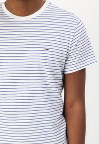 Hvit t-skjorte med horisontale blå striper, rund hals, og liten brodert logo i rød, hvit og blå på venstre bryst.