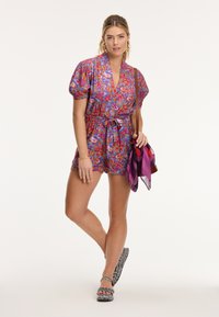 Színes mintás overall rövid puffujjakkal, V-nyakkivágással és összehúzott derékkal. Kicsi oldaltáskával és mintás cipőkkel viselve.