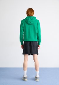 Green hoodie with a fitted design and front pocket, paired with black shorts and white crew socks. Features gray and white sneakers.