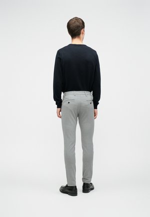 Homme portant une chemise noire à manches longues, un pantalon slim gris clair et des chaussures noires, debout dos à la caméra sur un fond uni.
