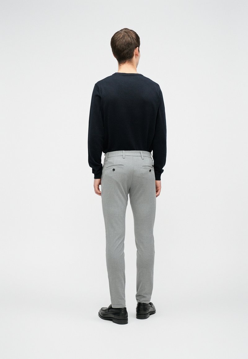 Homme portant une chemise noire à manches longues, un pantalon slim gris clair et des chaussures noires, debout dos à la caméra sur un fond uni.