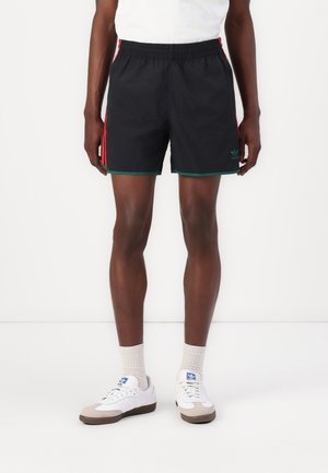 Personne portant un short de sport noir avec des bandes rouges sur les côtés, des chaussettes blanches et des baskets blanches sur un fond blanc uni.