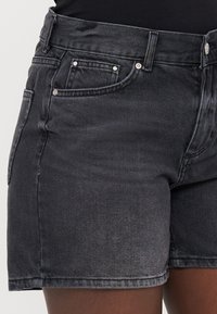 Shorts de mezclilla negra con un diseño clásico, cinco bolsillos y detalles en metal plateado. La tela tiene una ligera textura con un acabado desgastado.