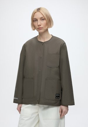 Frau mit kurzen blonden Haaren, die eine olivgrüne, geknöpfte Jacke mit drei Vordertaschen und weiße Hosen trägt, steht vor einem schlichten Hintergrund.