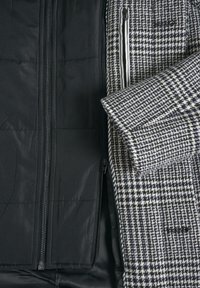 Schwarzes Innenfutter mit gesteppter Textur neben einer Außenschicht mit Hahnentrittmuster in Grau und Weiß, ausgestattet mit einem vorderen Reißverschluss.