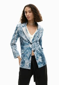 Blazer con motivo blu e bianco con maniche lunghe, rever a punte, chiusura con un bottone e tasche frontali. Indossato sopra una maglietta bianca.