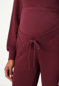 Burgundské tehotenské jogger súprava s jemnou textúrou, elastickým pásom a nastaviteľným šnúrkou. Obsahuje dlhé rukávy a priliehavý strih.