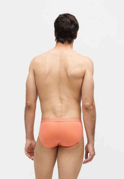 Άνδρας που στέκεται γυρισμένος με την πλάτη στην κάμερα, φορώντας εσώρουχο τύπου σλιπ σε κοραλλί χρώμα, με απλό φωτεινό φόντο.