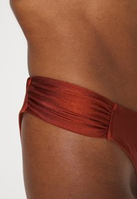 Pele castanha texturizada com um close-up de um biquíni castanho-avermelhado apresentando uma cintura larga e franzida e um design de cobertura mínima.