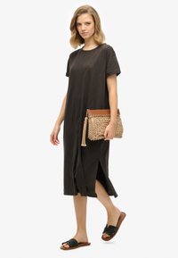 Robe grise foncée, longueur genou, avec une coupe ample, des manches retroussées et des fentes sur les côtés. Associée à une pochette tissée beige et des sandales noires à glissière.