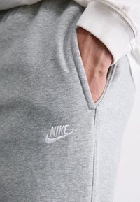Pelēki kokvilnas joggeri ar gludu tekstūru, ar sānu kabatām un baltu izšūtu Nike logo uz kreisā augšstilba. Baltas ribotas piedurknes.