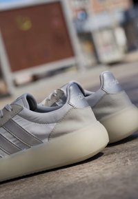 Paire de baskets Adidas gris clair avec semelles blanches placées à l'extérieur sur une surface en béton, montrant une vue arrière et latérale.