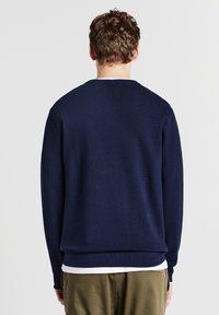Maglione lavorato a maglia blu navy con colletto rotondo, polsini e orlo a costine. Indossato su una maglietta bianca e abbinato a pantaloni verde oliva.