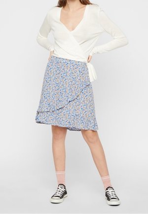 Weißes langärmeliges Wickeloberteil mit tiefem V-Ausschnitt, kombiniert mit einem blauen Minirock mit floralen Mustern und Rüschensaum sowie schwarzen Sneakern.