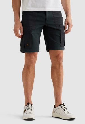 Mężczyzna w czarnych spodenkach typu cargo i białych trampkach, stojący z rękami swobodnie opuszczonymi wzdłuż ciała na jasno szarym tle.