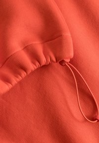 Koraaloranje stof met een zachte textuur, voorzien van gerimpelde details en een trekkoord met een ronde stopper voor een verstelbare pasvorm.