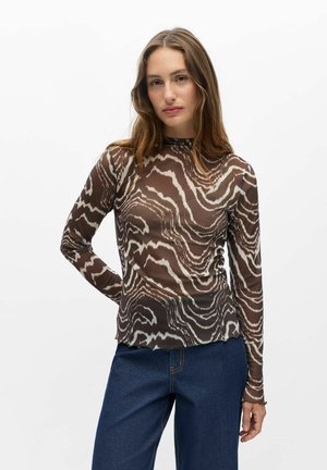 Femme aux longs cheveux bruns portant un haut moulant à manches longues avec un motif ondulé marron et blanc, et un jean bleu, debout devant un fond blanc.