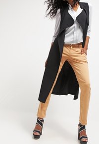 Gilet senza maniche nero indossato sopra una blusa bianca, abbinato a pantaloni beige chiaro e sandali con tacco neri con cinturini bianchi.