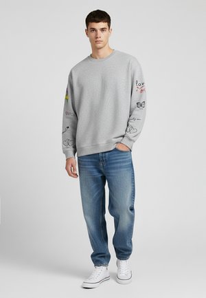 Sweatshirt gris avec des motifs graphiques sur les manches, associé à un jean bleu et des baskets blanches. Coupe confortable et design simple à col rond.