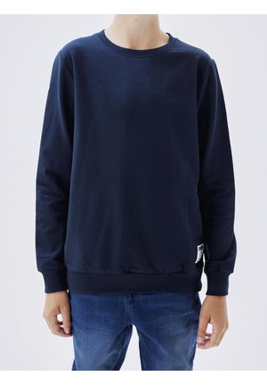 Enfant portant un sweat-shirt bleu marine uni et un jean bleu, debout contre un fond clair, les mains détendues le long du corps.