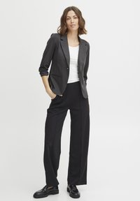 Svart skräddarsydd blazer med slag, kombinerad med lösa svarta byxor. Vit åtsittande linne underneath. Modellen står med händerna i fickorna.