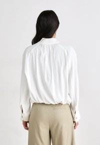White long-sleeve blouse with a gathered back yoke, soft texture, and buttoned cuffs; paired with light beige trousers.