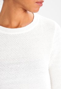 Vit stickad tröja med en texturerad, öppen vävdesign. Har en rund, ribbad halsringning och ett mjukt, andningsbart tyg.