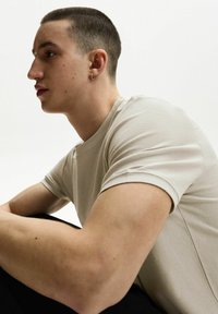 Svetle béžové krátké tričko s texturou a kulatým výstřihem, které nosí model sedící na jednobarevném pozadí.