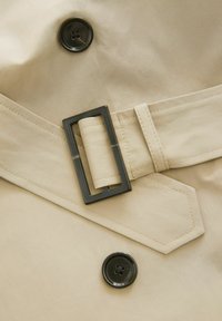 Un tissu de manteau trench beige avec un grand bouton noir et une boucle de ceinture rectangulaire. La ceinture présente des coutures contrastées et une texture lisse.