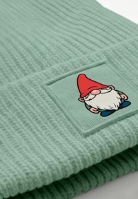 Mint green knit beanie with ribbed texture and a patch featuring a cartoon gnome with a red hat and white beard, surrounded by blue shoes.