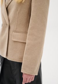 Beige blazer ze směsi vlny s klopovým límcem, jednou náprsní kapsou a zúženým střihem. Texturovaná tkanina a čisté linie zvýrazňují design.
