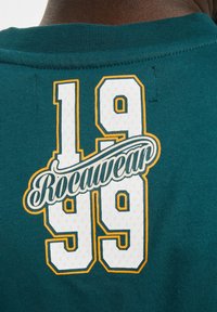 Zelená košile s krátkým rukávem a vroubkovaným výstřihem, na které se nachází bílé a oranžové logo "Rocawear" a nápis "1999" vytištěný na zádech.