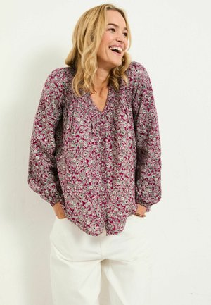 Fließende kastanienrote Bluse mit floralem Muster, V-Ausschnitt und langen Ärmeln. Weicher Stoff, gerafftes Detail am Ausschnitt, kombiniert mit hellen Hosen.