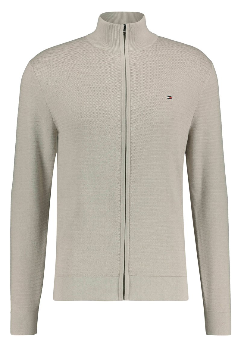 Gilet zippé gris clair en tissu tricoté texturé, avec un col haut et des poignets côtelés. Petit logo sur le côté gauche de la poitrine.