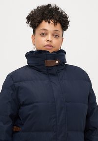 Tumesinine puffer-jakk kõrge kraega, millel on pruun nahkdetail ja pehme karvane äär, loodud soojuse ja mugavuse tagamiseks.