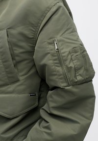 Zaļa pūkaina jaka ar teksturētu audumu, kurai ir rāvējslēdzēja kabata uz piedurknes un uzliekamā kabata priekšpusē, redzama zīmola etiķete.