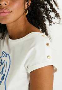 Camiseta de algodón blanca con bordado azul, mangas enrolladas y botones dorados en el hombro. El modelo tiene el cabello rizado y un maquillaje mínimo.