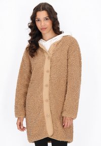 Cappotto teddy in pelliccia sintetica color sabbia con un davanti button-up beige liscio, vestibilità oversize e maniche lunghe. L'interno presenta un cappuccio bianco.