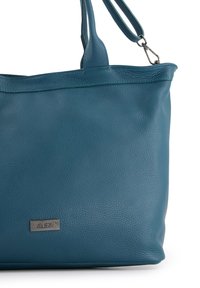 Teal læder tote taske med en struktureret overflade, der har en sølvfarvet logo-plade, to skulderstropper og et minimalistisk design.