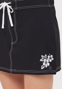 Falda de baño negra con costuras blancas, que presenta un estampado floral en la esquina inferior izquierda y una cintura con cordón.