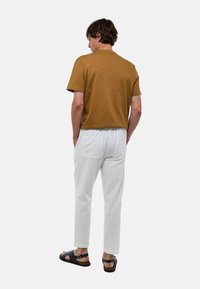 Chemise marron à manches courtes, pantalons blancs texturés légers avec taille élastique, et sandales noires. Poche arrière visible sur le pantalon.