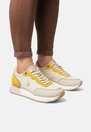 Persona che indossa sneaker beige e gialle con lacci, pantaloni marroni risvoltati su sfondo neutro.