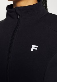 Gros plan sur une personne portant une veste noire à fermeture éclair avec un col montant et un logo "F" stylisé blanc sur la poitrine.