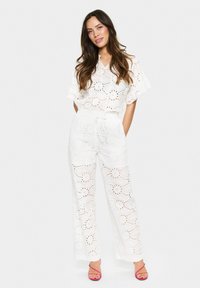 Witte tweedelige outfit met geborduurde bloemendetails, een top met korte mouwen en een broek met wijde pijpen en een trekkoord in de taille, gecombineerd met roze sandalen.