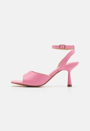 Sandale à talons hauts rose avec une sangle de orteil matelassée, une sangle fine autour de la cheville avec une boucle dorée, et un talon aigu courbé sur fond blanc.