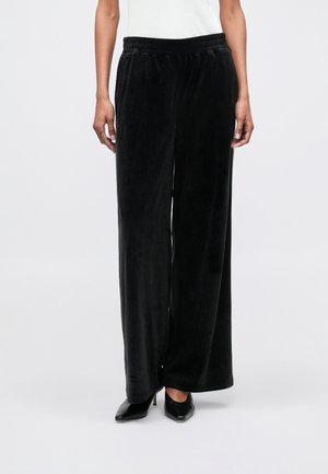 Pantalons noirs à jambes larges en tissu doux et texturé. Dotés d'une taille élastique pour le confort. Portés avec des talons noirs.