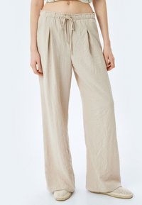 Beige wijde pantalon van gestructureerde stof, met een koord in de taille en voorste plooien. Gekleed met beige geweven instappers.