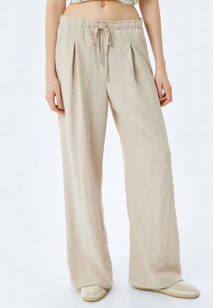 Beige weite Hosen aus strukturiertem Stoff, mit einem Kordelzugbund und vorderen Falten. Kombiniert mit beigen geflochtenen Slippern.