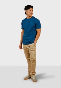 Camiseta de algodón azul con un pequeño logo, combinada con pantalones beige claros. El modelo lleva zapatillas grises con acentos en color marrón. Diseño simple y casual.