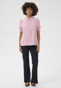 Ružová blúzka s golierom a naberanými rukávmi, so zapínaním na gombíky vpredu a textúrovanými detailmi. V kombinácii s tmavými rozšírenými džínsami a obuvou so zvieracím motívom.